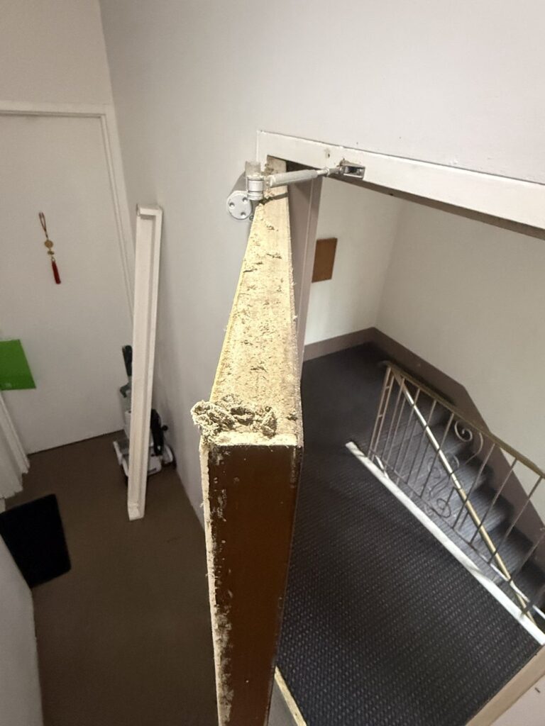 The top edge of a stairwell fire door with crumbling core material breaking away beside the closer